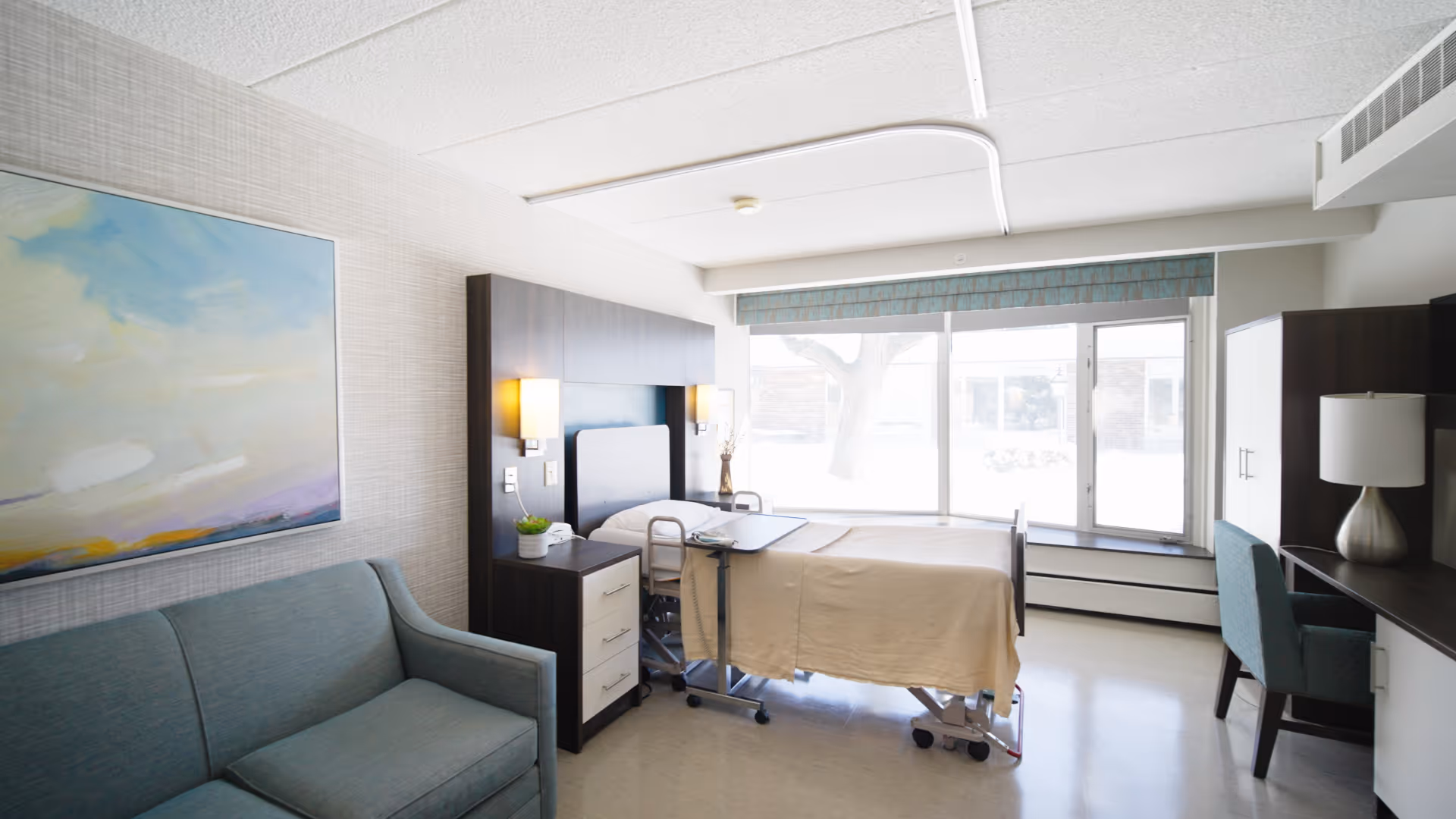 A bright and clean nursing facility room with a hospital bed covered in a beige blanket, a small bedside table with a plant, a teal sofa, a large window with a view outside, a desk with a chair, and a lamp on a cabinet.
