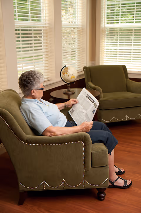 An elderly woman with short gray hair and glasses is sitting in a green upholstered armchair reading a newspaper. The room has large windows with white blinds, wooden flooring, and another matching green armchair next to a small round table with a globe on it.