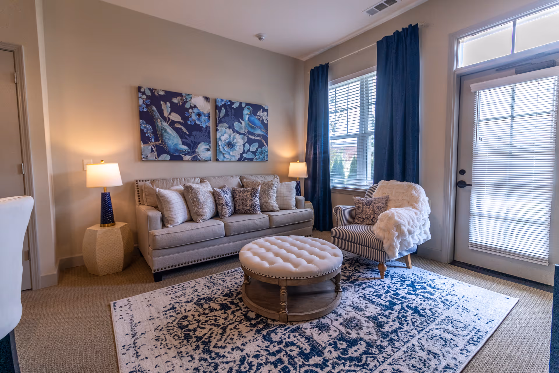 A cozy living room with a beige sofa adorned with multiple decorative pillows, a round tufted ottoman coffee table, a striped armchair with a white throw blanket, two table lamps on either side of the sofa, blue curtains framing a window, and a glass door with blinds.