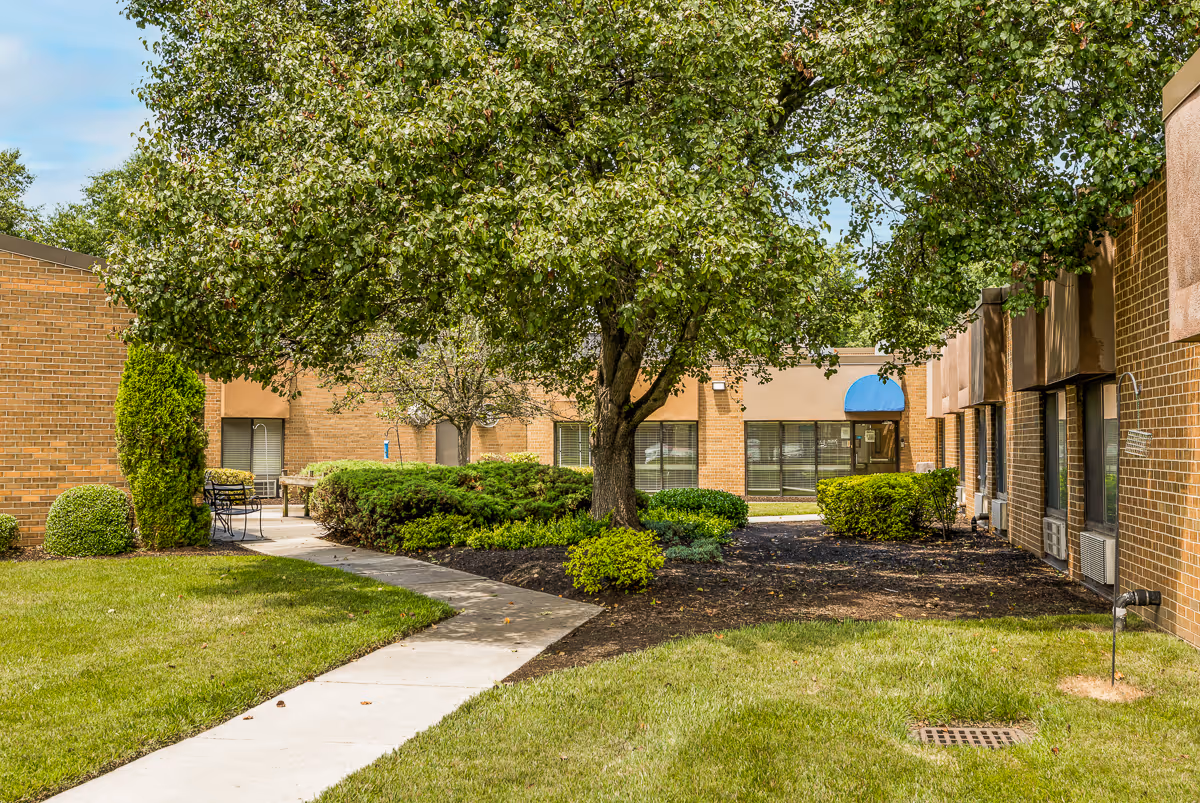 Outdoor courtyard area at Autumn Lake Healthcare at Patuxent River featuring a concrete walkway, green grass, trimmed bushes, a large leafy tree, and brick building walls with windows and a blue awning.