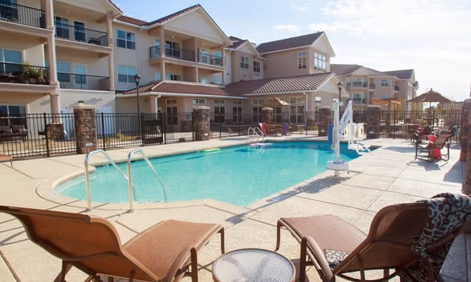 Outdoor swimming pool area at Estrella Estates Gracious Retirement Living with lounge chairs, a small round table, and a multi-story residential building in the background under a clear sky.