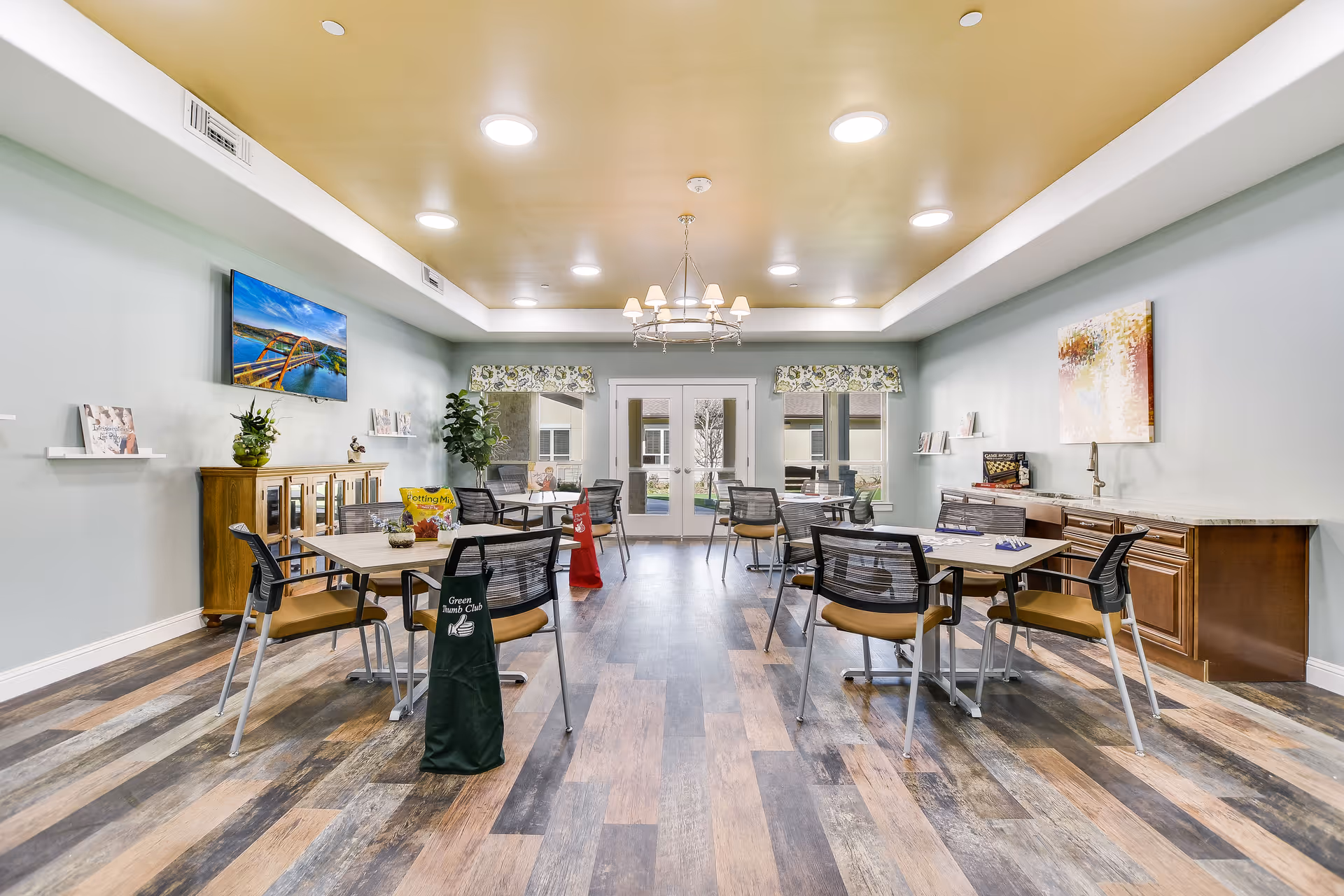 A bright and spacious common activity room with several tables and chairs arranged for group activities. The room features a wooden floor with varied tones, a yellow ceiling with recessed lighting and a chandelier, light blue walls, and large windows with floral valances letting in natural light. There is a TV mounted on the wall, a wooden cabinet with plants and books, and a countertop with cabinets along one side. Some tables have game pieces and bags labeled 'Green Thumb Club' and 'Potting Mix'.
