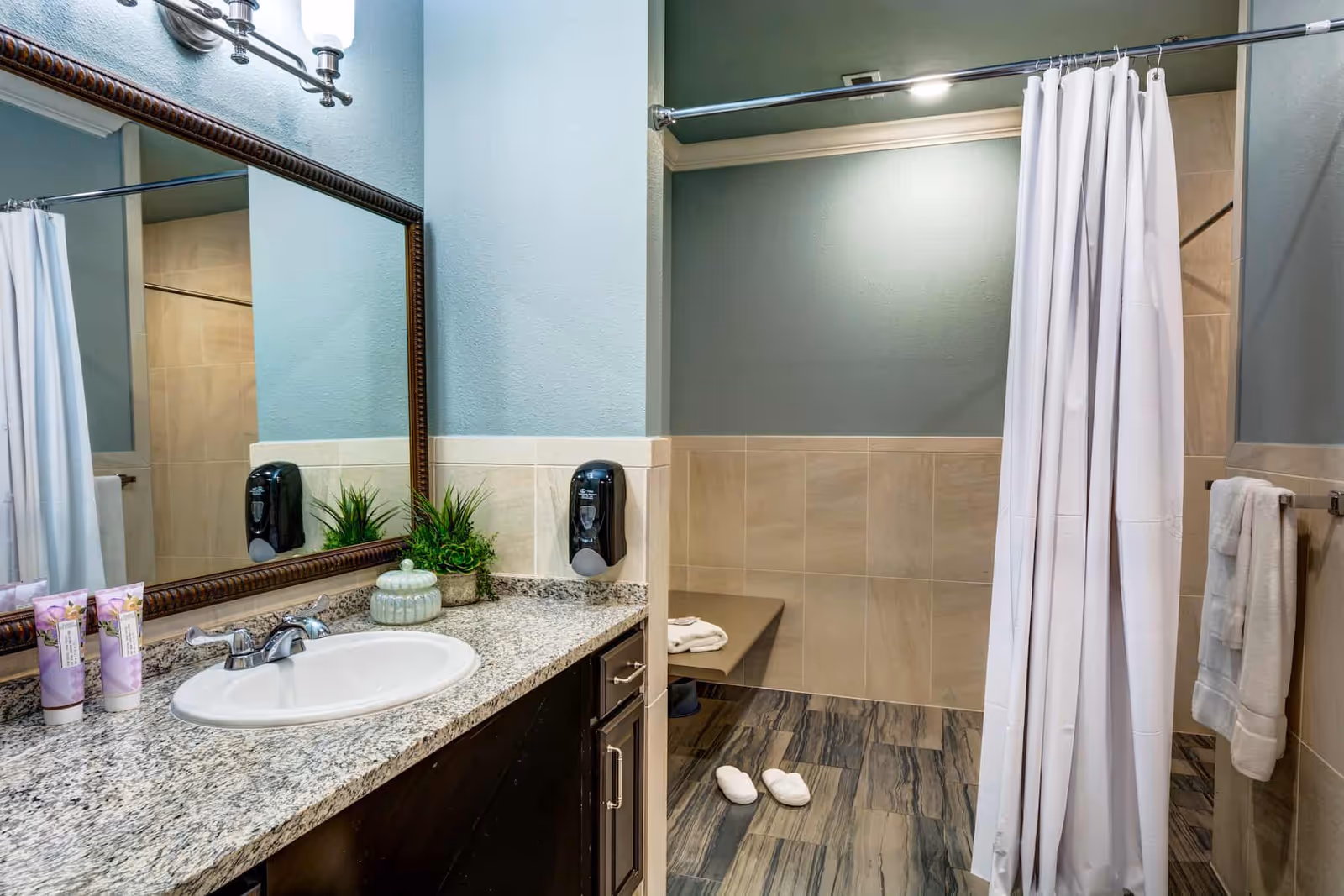 A clean and modern bathroom with a granite countertop sink, a large framed mirror, and a shower area with a white curtain. The shower area has a built-in bench, beige tiled walls, and a pair of white slippers on the floor. There are hand towels hanging on a rack and a soap dispenser mounted on the wall.