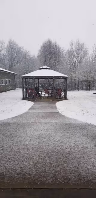 Snow-covered outdoor pathway leading to a gazebo with chairs inside, surrounded by snow-covered ground and leafless trees in the background during snowfall.