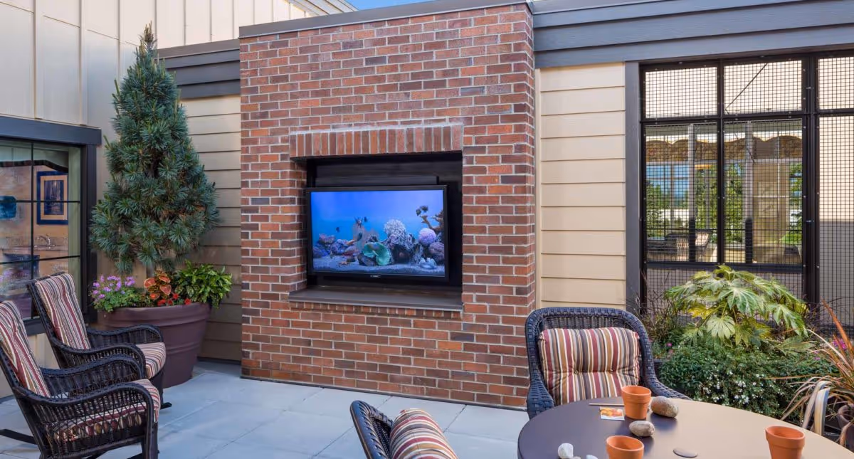 Outdoor patio area with a brick fireplace that has a TV mounted inside it. There are several wicker chairs with striped cushions around a round table with small flower pots and stones. Potted plants and greenery surround the seating area, and there is a window and a door with a mesh screen in the background.