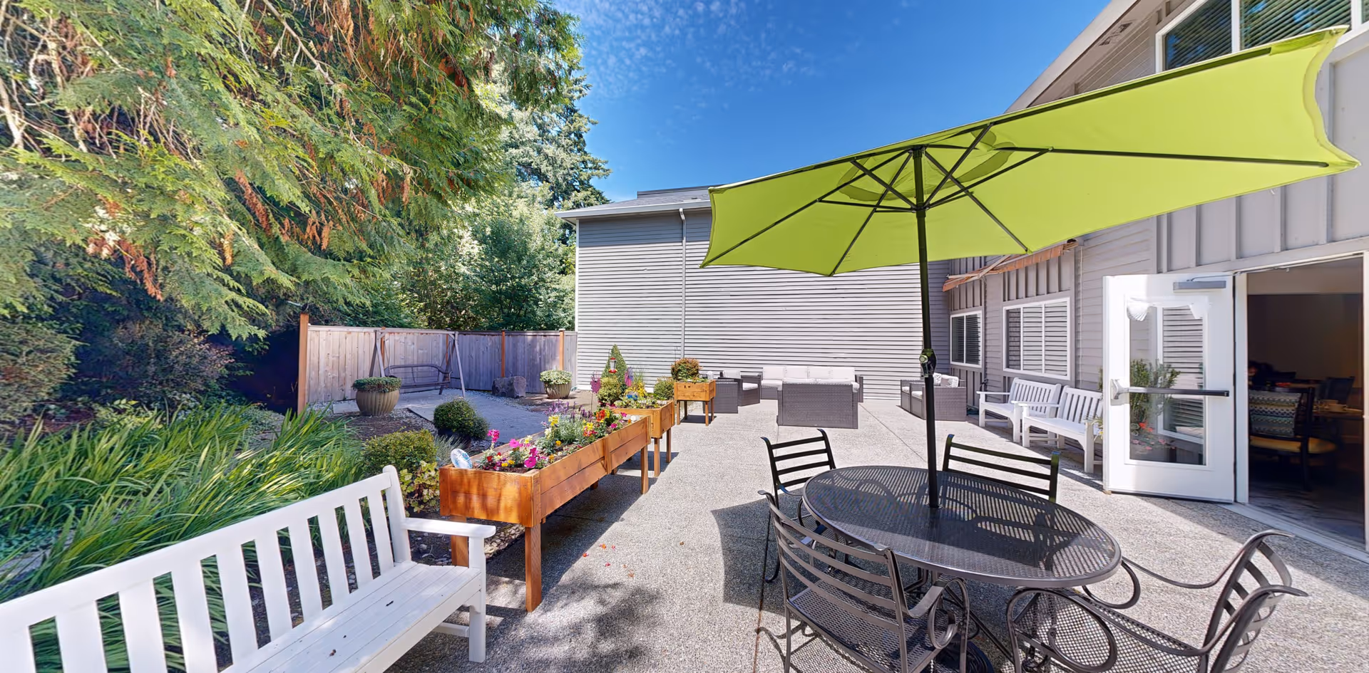 Sunny outdoor courtyard patio with a round table, chairs under a green umbrella, benches, and raised planters next to the building.