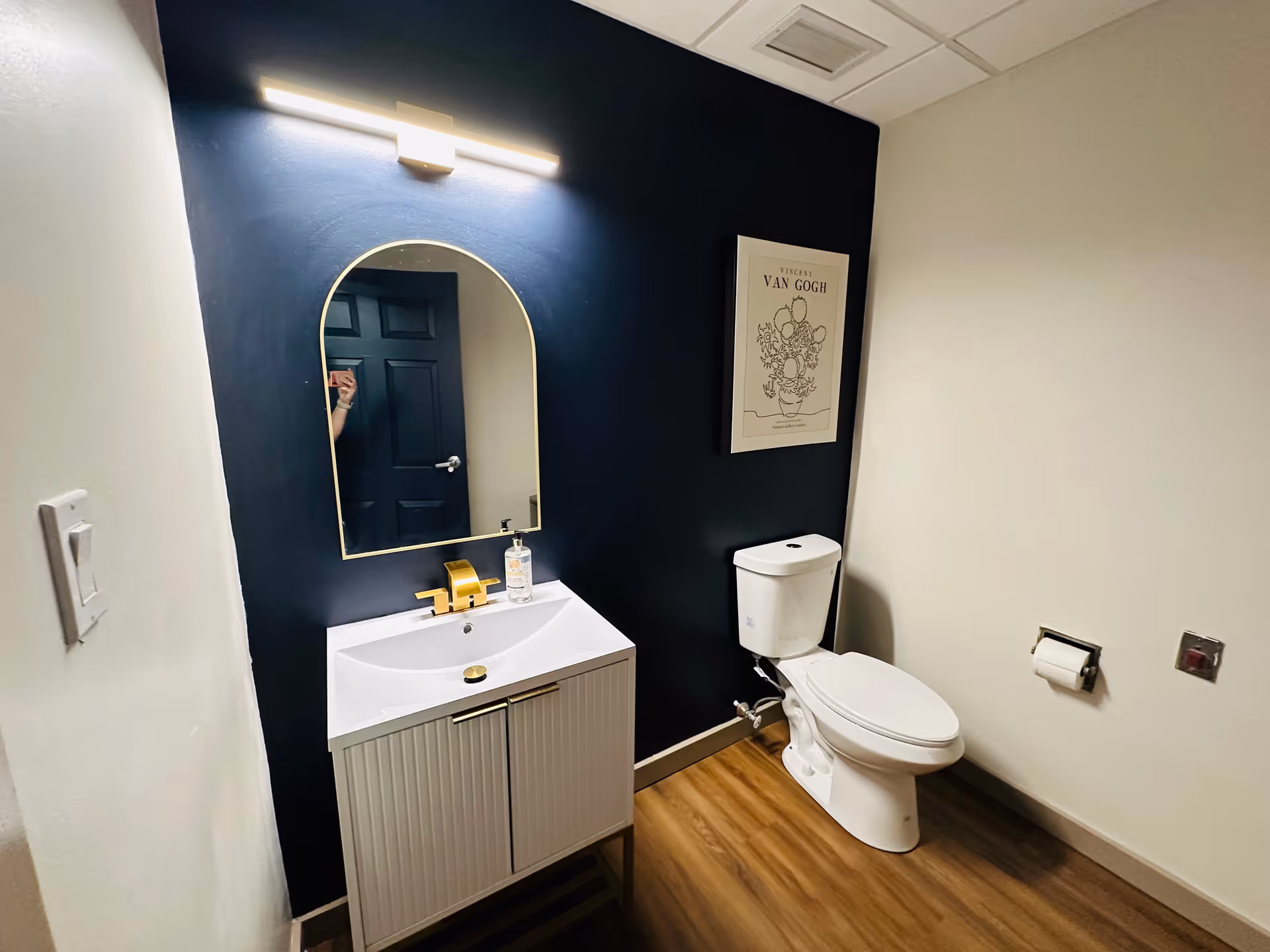 A modern bathroom with a white toilet and a white vanity with a gold faucet. Above the vanity is an arched mirror with a light fixture above it. The wall behind the toilet and vanity is painted dark blue, and there is a framed Vincent Van Gogh print hanging on the wall. The floor is wood-style laminate.