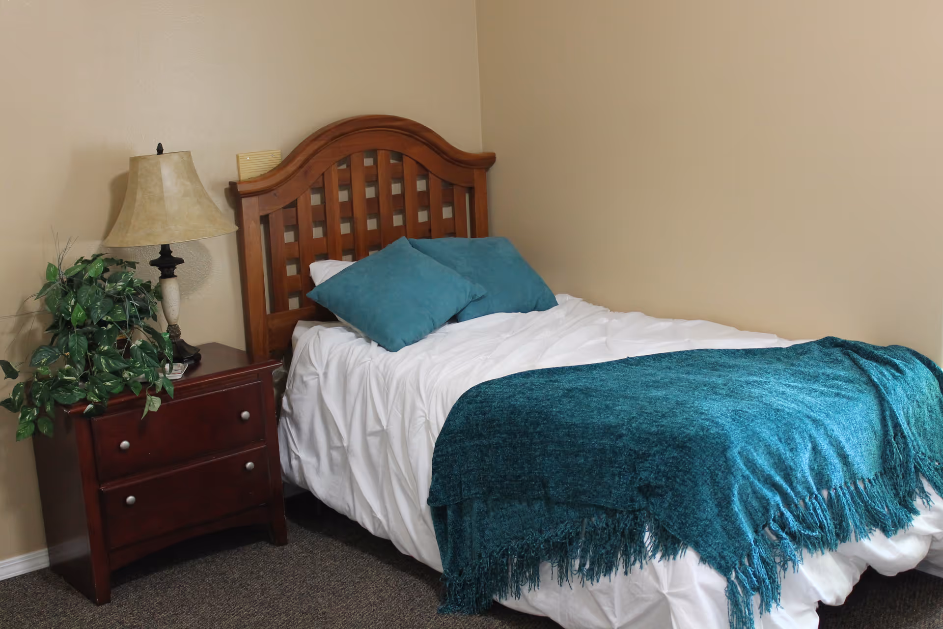 A small bedroom with a single wooden bed featuring a lattice headboard. The bed is made with white bedding, two teal pillows, and a teal throw blanket. Next to the bed is a dark wooden nightstand with two drawers, a beige lamp, and a green leafy plant. The walls are beige and the floor is carpeted in a dark color.