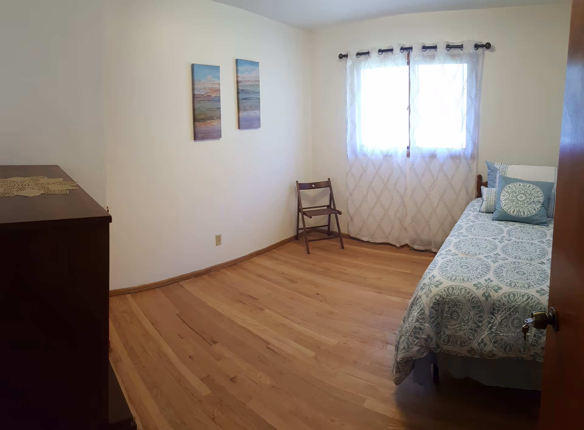 A simple bedroom with wooden flooring, a single bed with patterned blue and white bedding and pillows, a wooden chair near a window with sheer white curtains, two landscape paintings on the wall, and a wooden dresser with a lace doily on top.