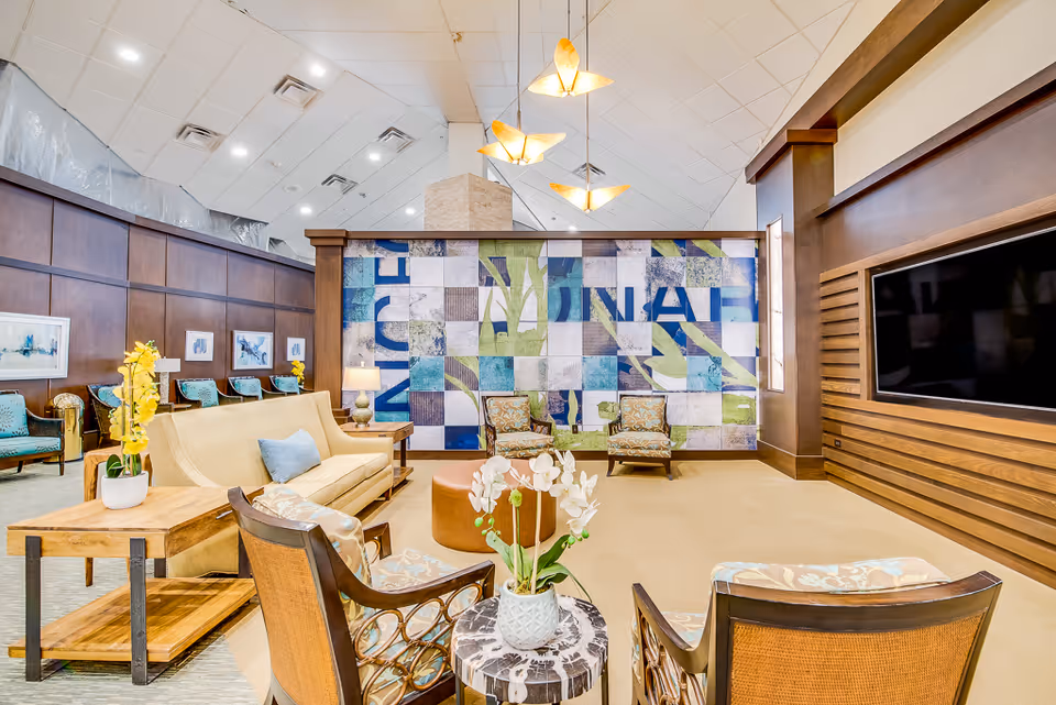 A spacious and well-lit living room area with a high ceiling and modern pendant lights. The room features a beige sofa with a blue pillow, several patterned armchairs, wooden side tables with decorative plants, and a large wall-mounted flat-screen TV. The back wall has a colorful abstract tiled mural with blue, green, and white tones. The walls are paneled with dark wood, and there are framed artworks along one side.