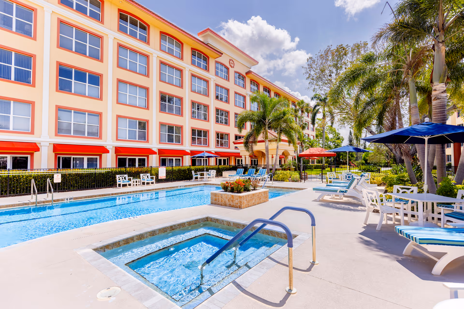 Outdoor pool and hot tub with lounge chairs and umbrellas next to a multi-story residential building and palm trees.