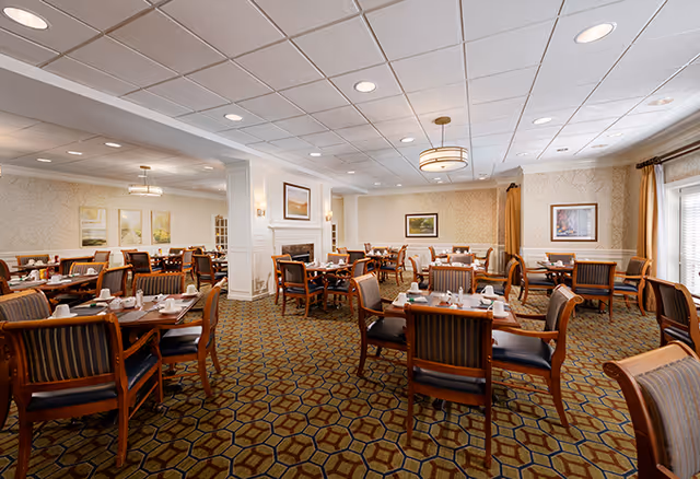 A spacious dining room in a senior living facility with multiple wooden tables and chairs arranged neatly. Each table is set with white cups, plates, and napkins. The room features patterned carpet flooring, beige wallpaper, framed artwork on the walls, and ceiling lights. Large windows with curtains allow natural light to enter the room.