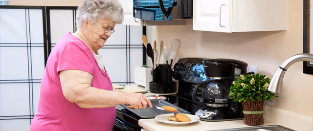 An elderly woman in a pink shirt is cooking in a kitchen, placing cookies on a plate on the countertop. The kitchen has a stove, coffee maker, microwave, utensils in a holder, and a small potted plant near the sink.