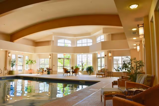 Indoor swimming pool area with large windows letting in natural light, surrounded by wicker chairs with cushions, small tables, and potted plants. The ceiling features wooden beams and the space has a warm, inviting atmosphere.