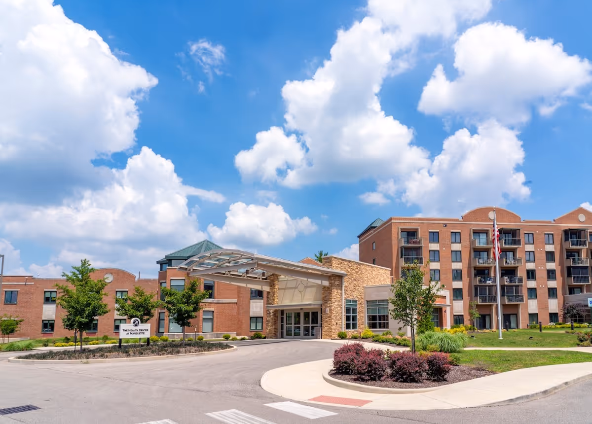 Exterior view of a large brick building with multiple floors and balconies under a blue sky with scattered clouds. The building has a covered entrance with a glass and metal canopy. There are small trees and landscaped bushes around the driveway leading to the entrance. An American flag is visible on a flagpole near the building.
