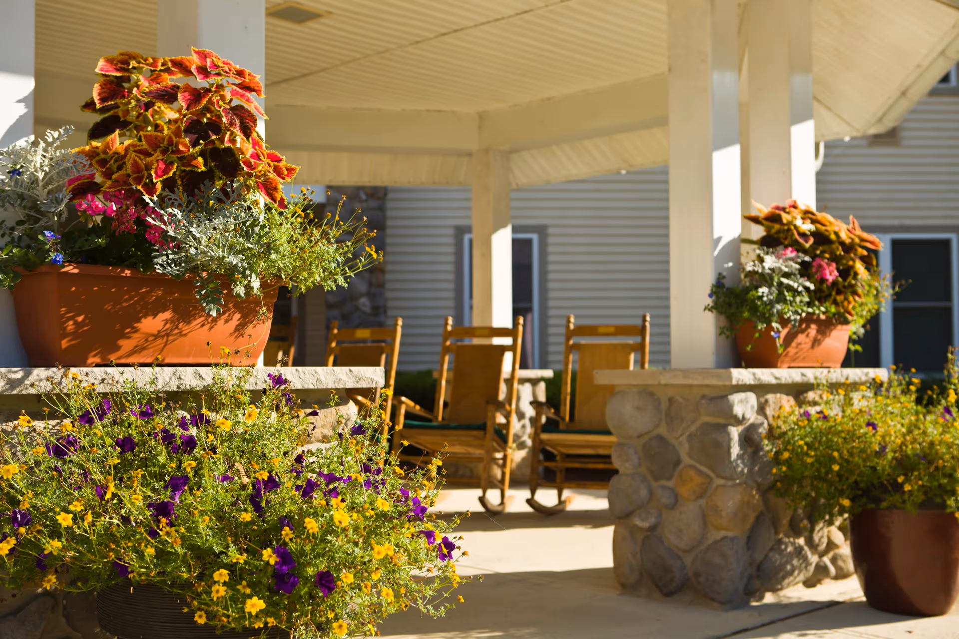 A shaded outdoor patio area with stone pillars and wooden rocking chairs, surrounded by vibrant flower pots containing colorful flowers and foliage plants.