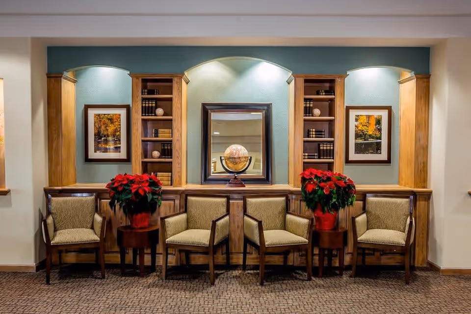 A cozy seating area with four cushioned wooden chairs arranged in a row. Between the chairs are two small round tables each holding a red poinsettia plant. Behind the chairs is a wooden built-in shelving unit with books, decorative spheres, and a globe in front of a mirror. The wall is painted light blue with two framed autumn landscape pictures on either side of the shelving unit. The floor is carpeted with a patterned design.