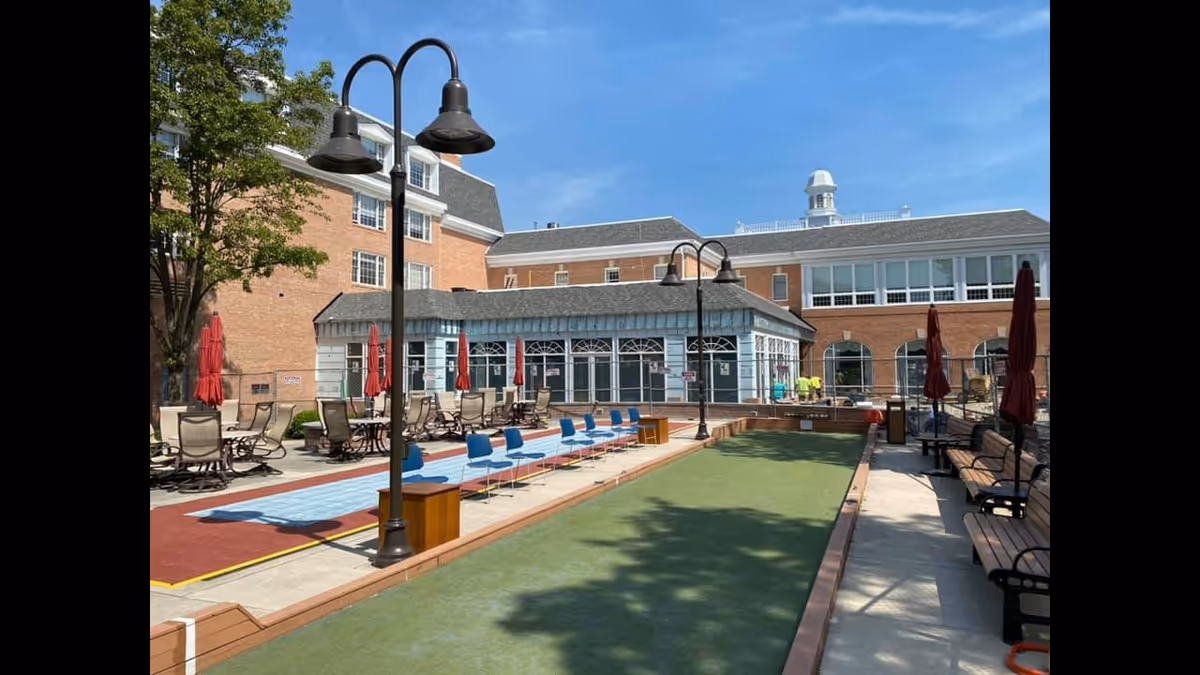 Outdoor courtyard area at Riddle Village LifeCare Retirement Community featuring a bocce ball court, shuffleboard court with blue chairs lined up along its side, several benches, patio tables with umbrellas, and a multi-story brick building in the background under a clear blue sky.