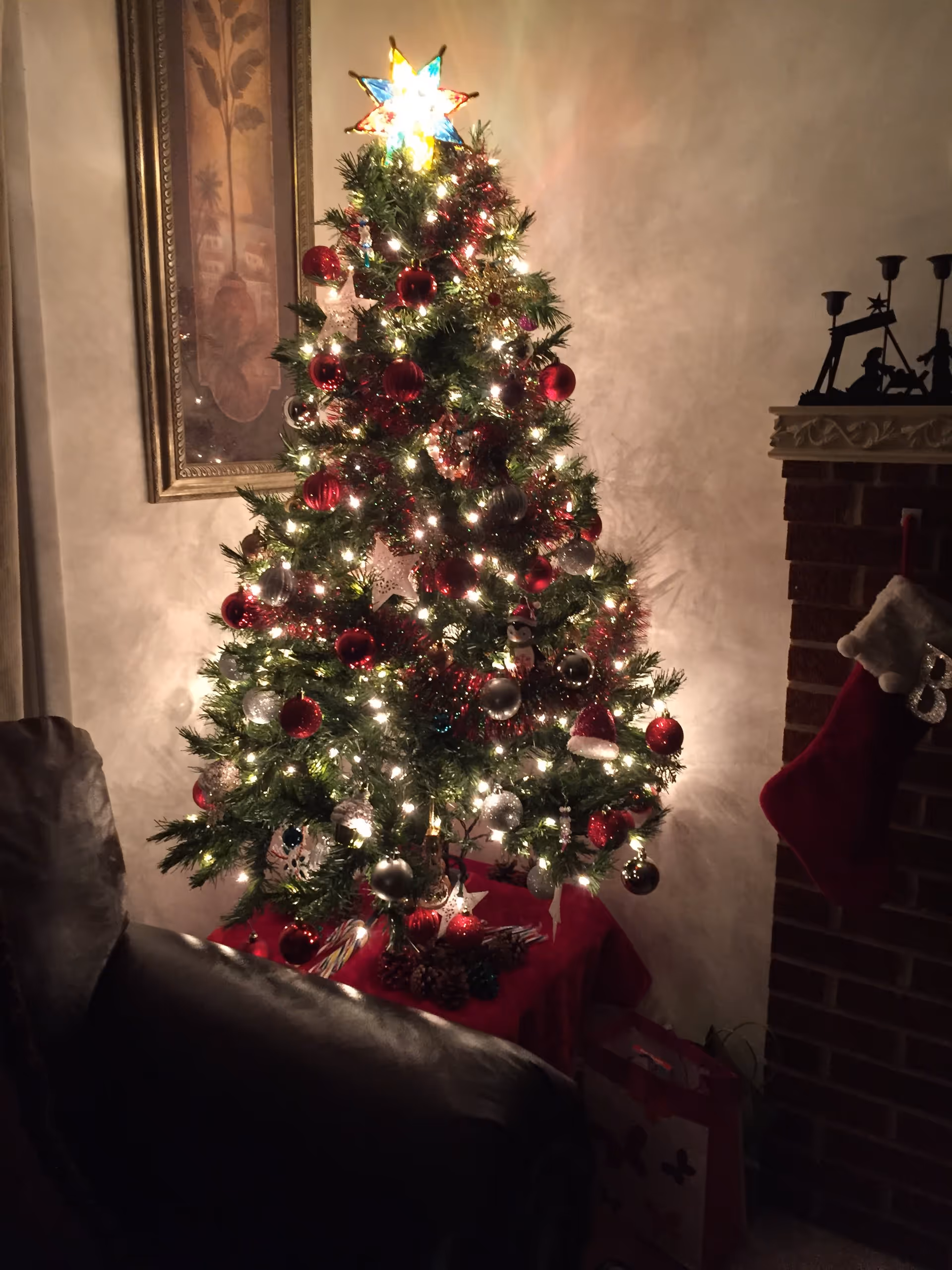 A decorated Christmas tree lit with white lights and red and silver ornaments standing next to a brick fireplace with a stocking.