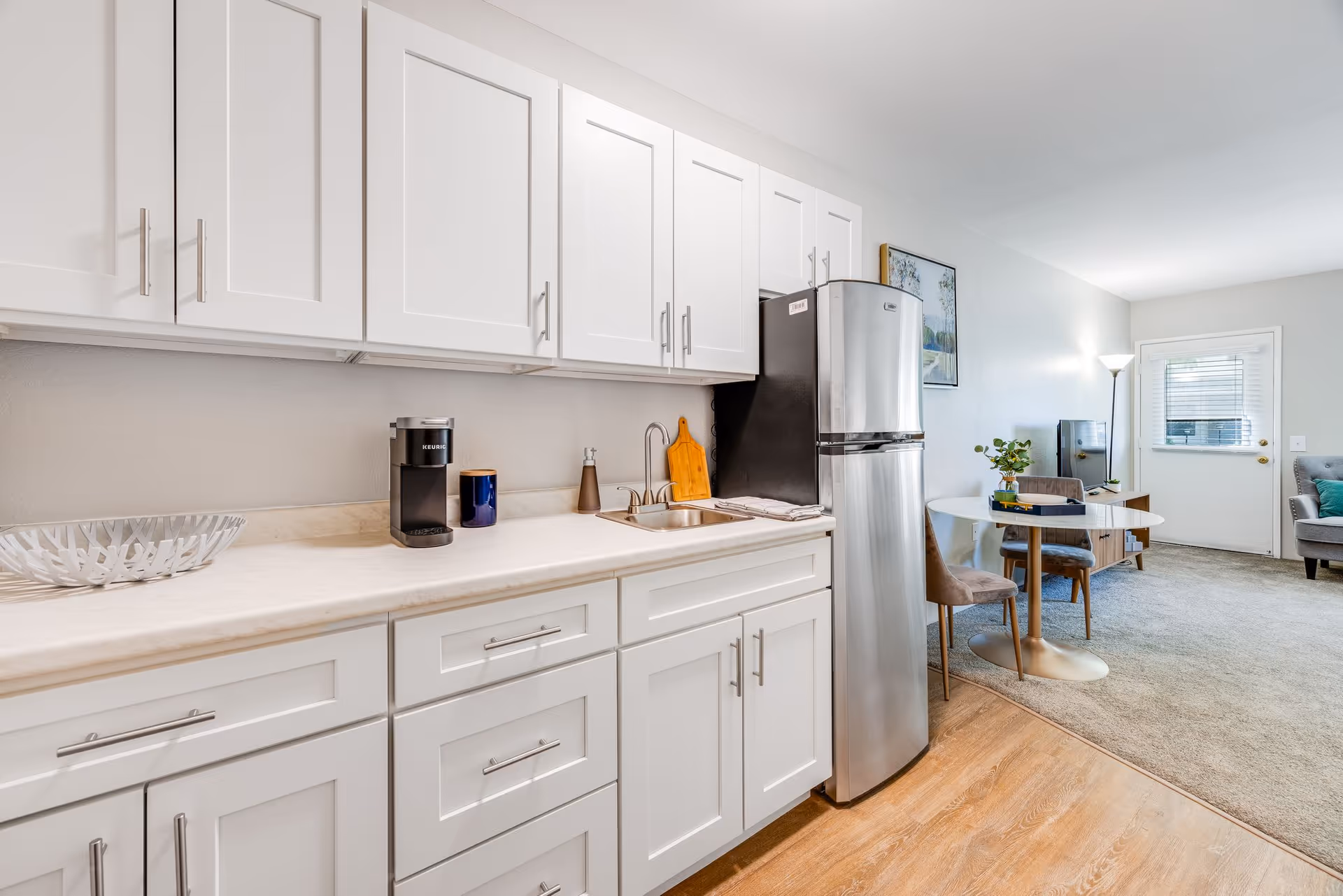 A modern kitchen area with white cabinets, a stainless steel refrigerator, a small sink, and a coffee maker on the countertop. Adjacent to the kitchen is a small dining area with a round table and two chairs, and a living room space with a gray armchair, a floor lamp, and a door with a window.