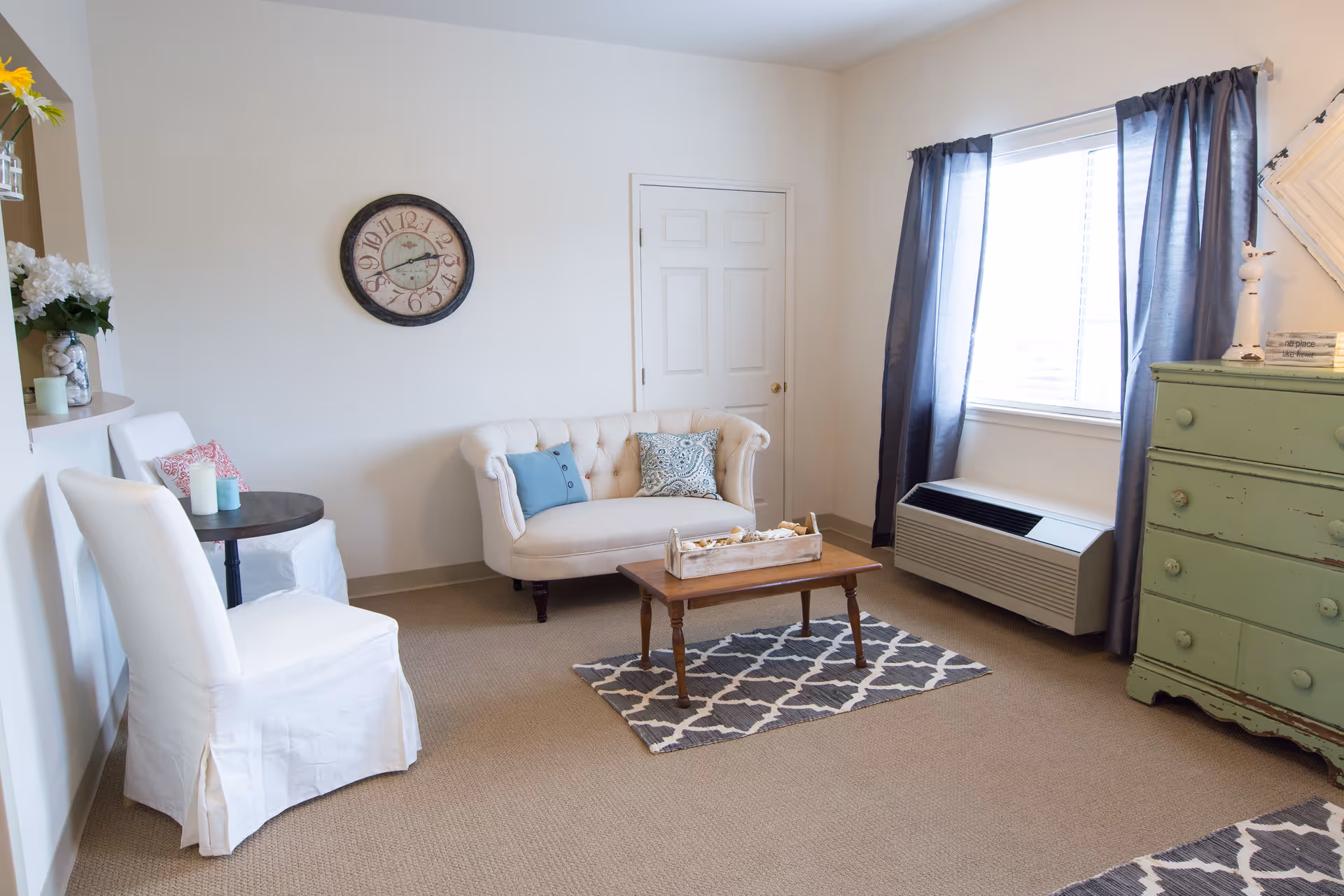 Sunlit living room with a small tufted loveseat, coffee table on a patterned rug, white chairs, and a green dresser by a window.