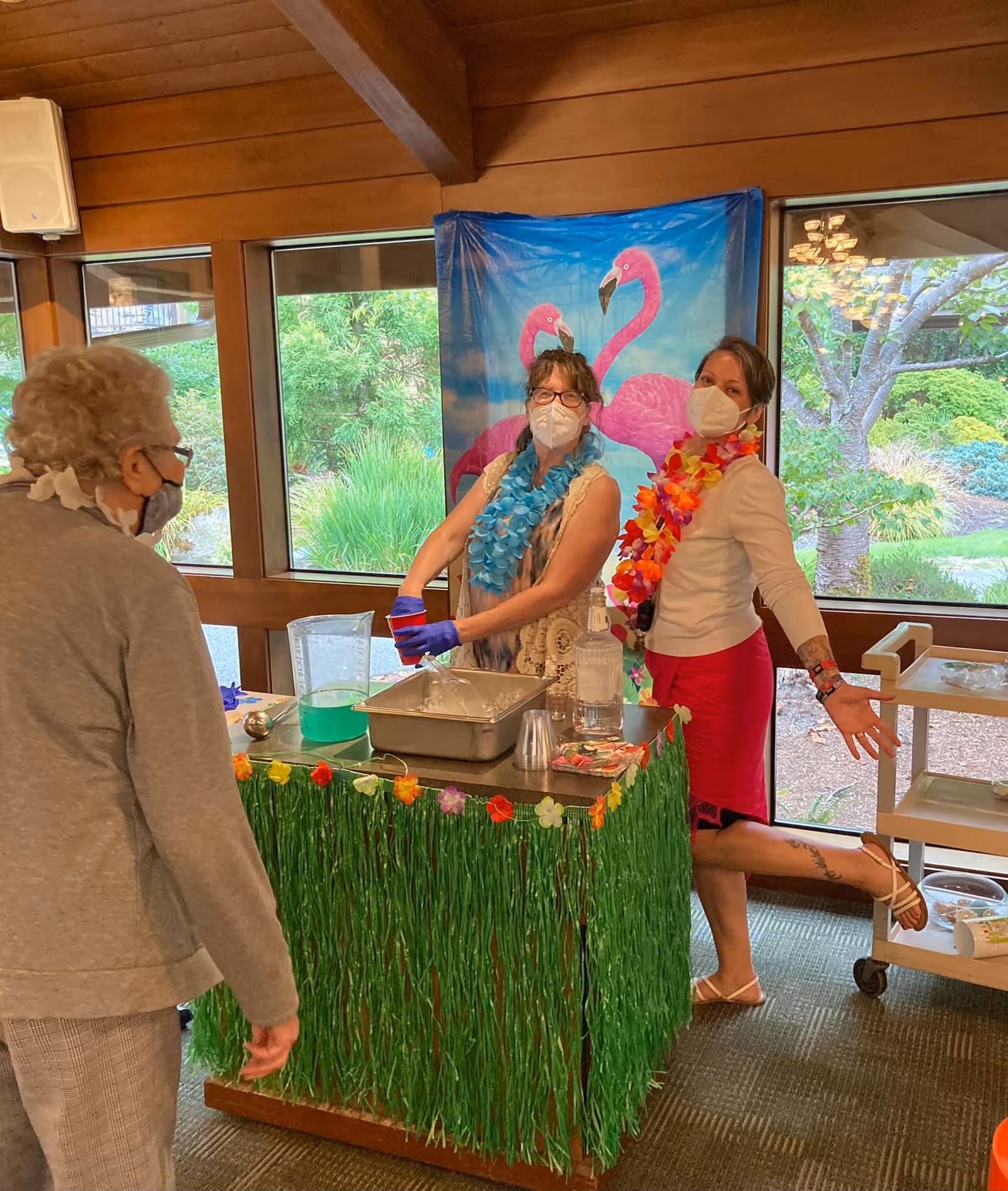 Two women wearing face masks and colorful leis stand behind a table decorated with green grass skirt and flowers, serving drinks to an elderly woman also wearing a mask. Behind them is a window showing greenery outside and a backdrop with two pink flamingos.
