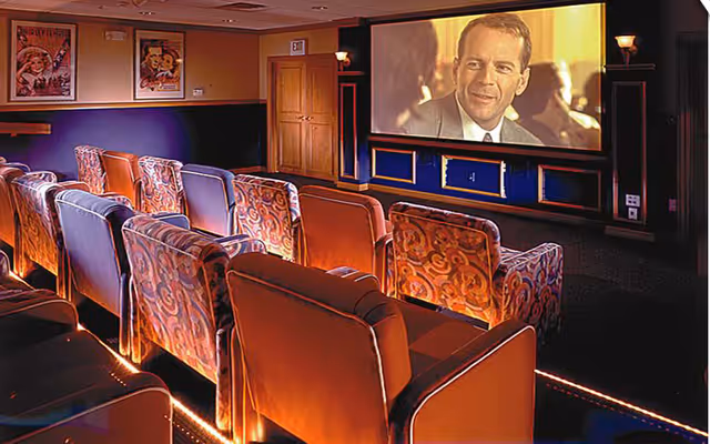 A cozy movie theater room with multiple rows of cushioned armchairs facing a large screen showing a man in a suit. The room has warm lighting, framed movie posters on the walls, and a door at the back.