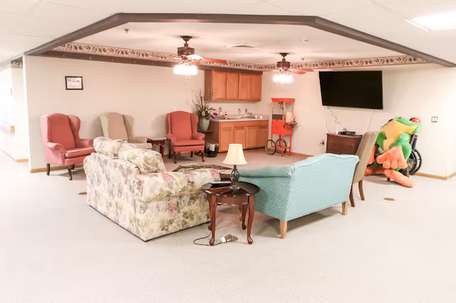 Communal living room area with sofas and armchairs arranged around side tables, a small kitchenette, wall-mounted TV, and a large plush frog toy.