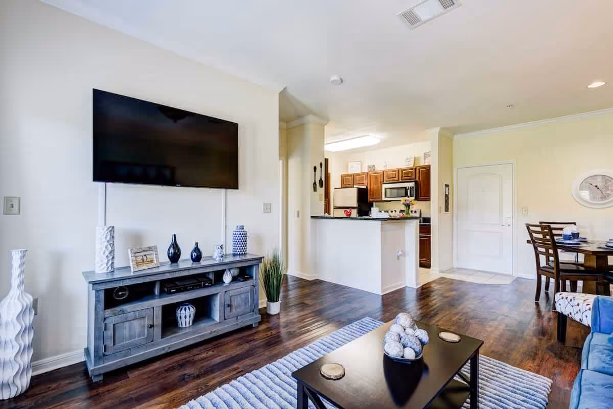 Bright and modern living area with a mounted flat-screen TV above a wooden console table decorated with vases and picture frames. The room features dark hardwood floors, a black coffee table with decorative balls, a blue couch, and a dining table with chairs in the background. The kitchen area with wooden cabinets, stainless steel appliances, and a white countertop is visible beyond the living space.