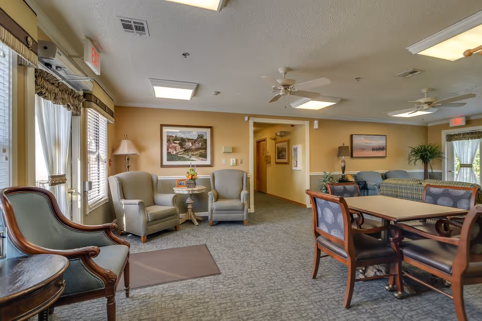 A cozy senior living facility common area with several upholstered armchairs and a table with four chairs. The room has beige walls, carpeted floors, ceiling fans, and framed artwork on the walls. Large windows with curtains allow natural light to enter the space.