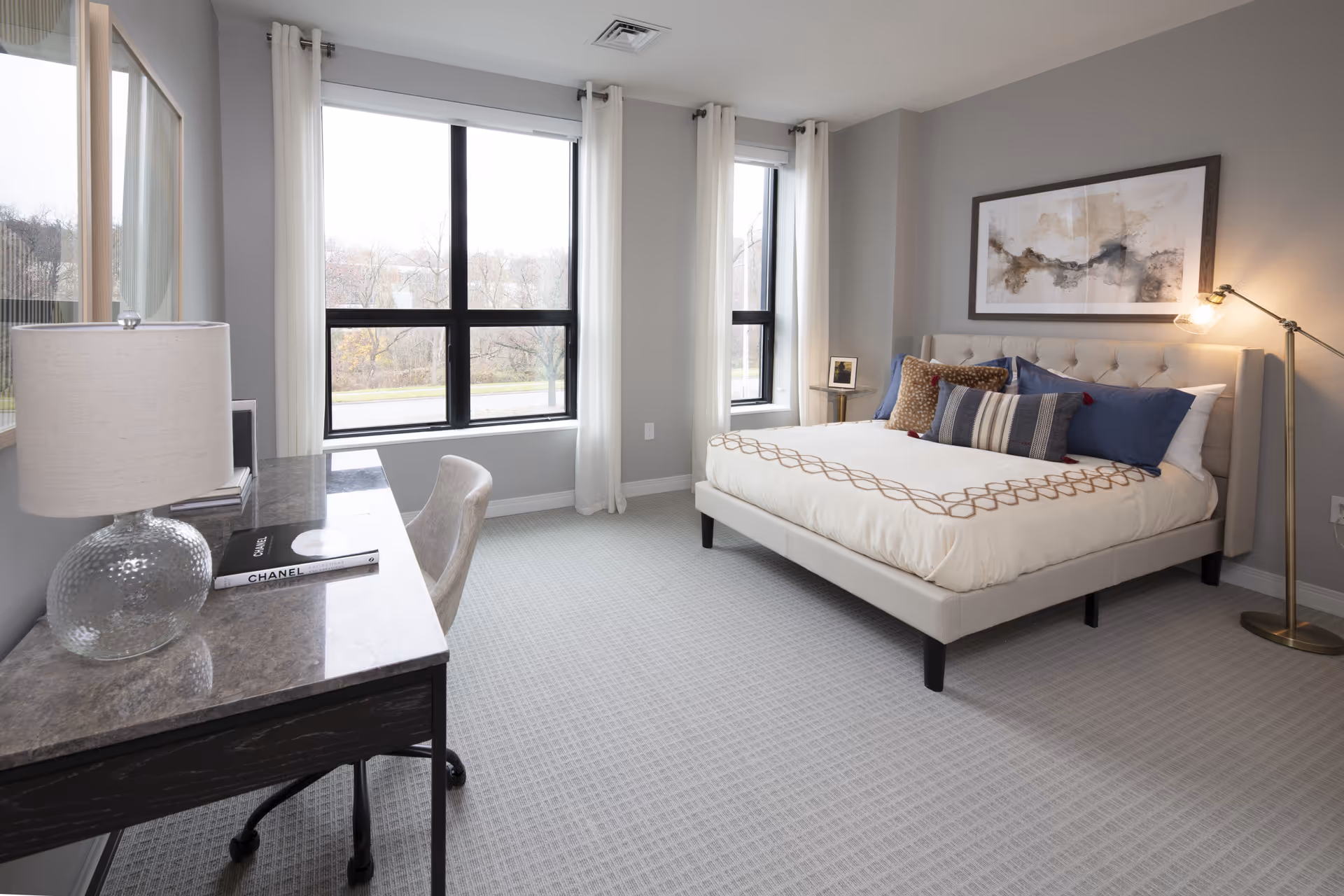 Bright, modern bedroom with a bed, desk, large windows, and neutral decor.