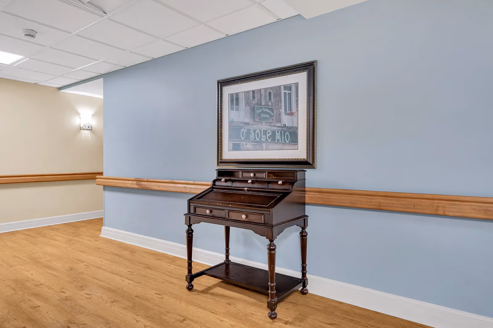 A hallway with a small dark wooden writing desk against a light blue wall beneath a framed picture and a wooden handrail.