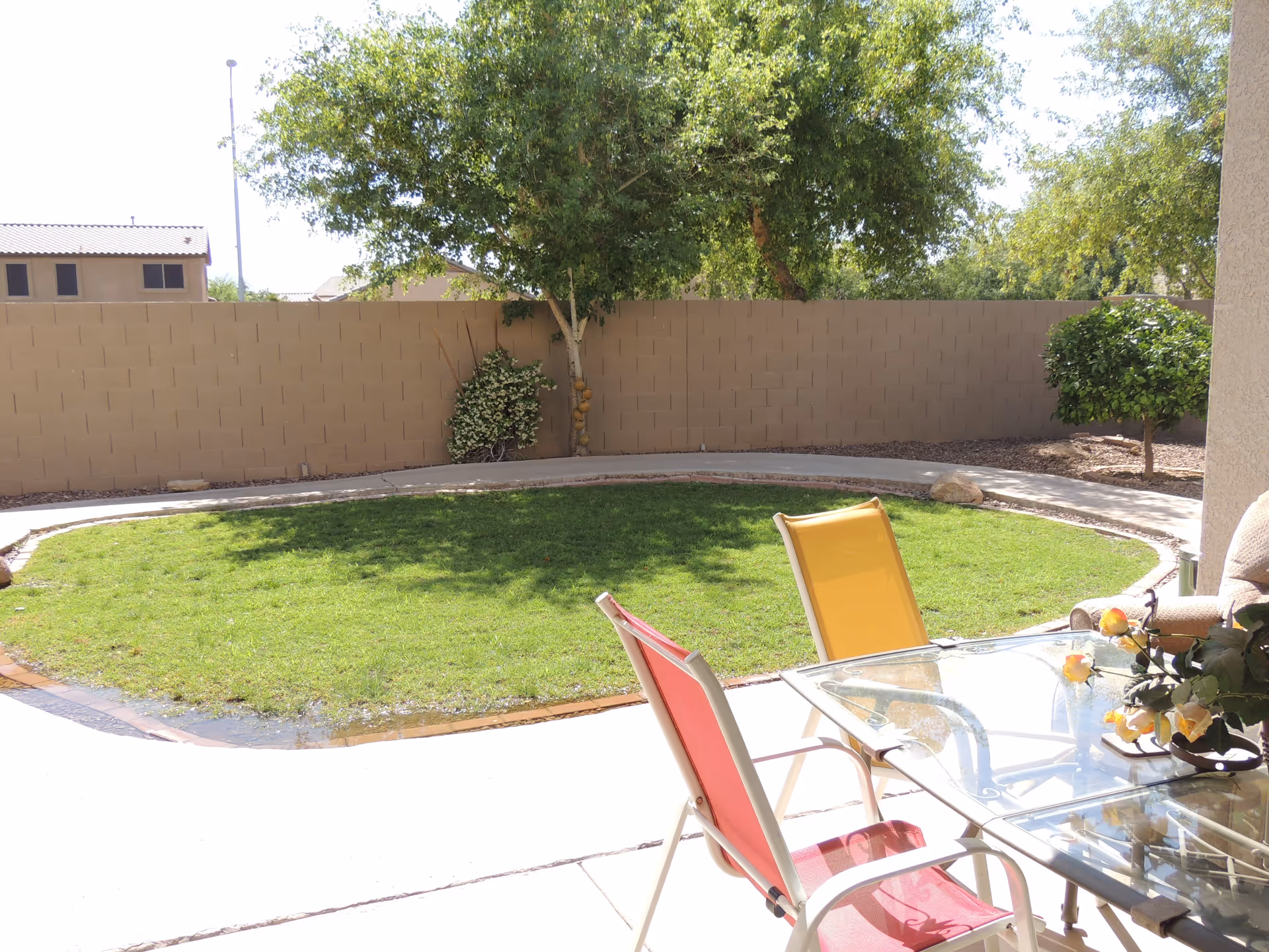 Patio with a glass table and colorful chairs overlooking a small circular lawn, trees, and a block privacy wall.