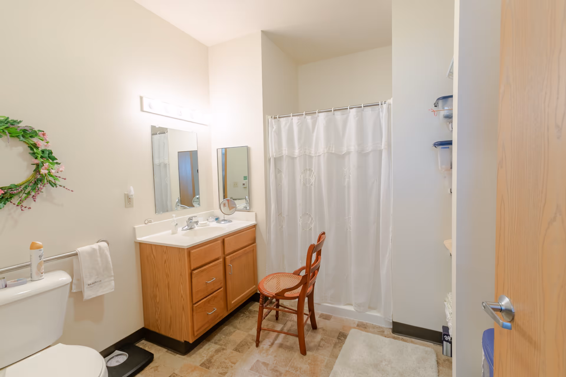 Well-lit bathroom with a vanity and mirror, toilet, shower with a white curtain, and a wooden chair.