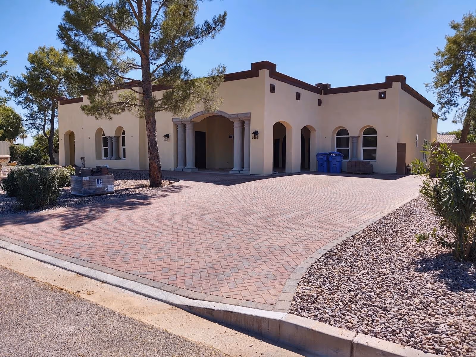Single-story stucco building with arched entryways, columns and a large paved brick driveway surrounded by gravel landscaping and trees.