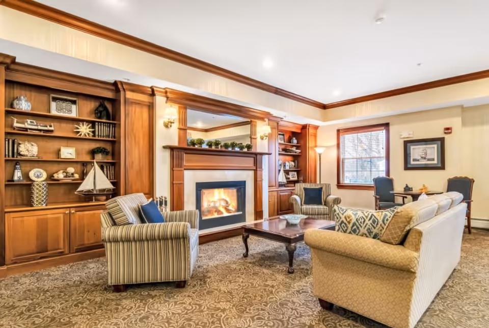 A cozy living room with a lit fireplace surrounded by wooden built-in shelves filled with decorative items. The room has a patterned carpet, a beige sofa with patterned cushions, two striped armchairs with blue pillows, a wooden coffee table, and a window with wooden blinds. There is also a small table with two chairs near the window and a framed picture on the wall.