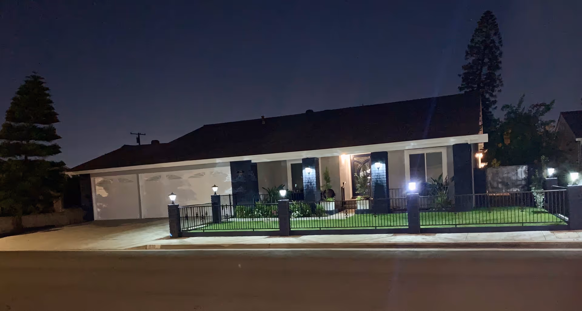 Front exterior of a single-story house at night with illuminated porch lights, a fenced lawn, and a two-car garage.