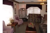 A cozy living room with multiple upholstered chairs and sofas arranged around a wooden coffee table with a floral centerpiece. There is a large birdcage in the corner near two windows with sheer white curtains and dark valances. The room has soft lighting from a ceiling fixture and a table lamp.
