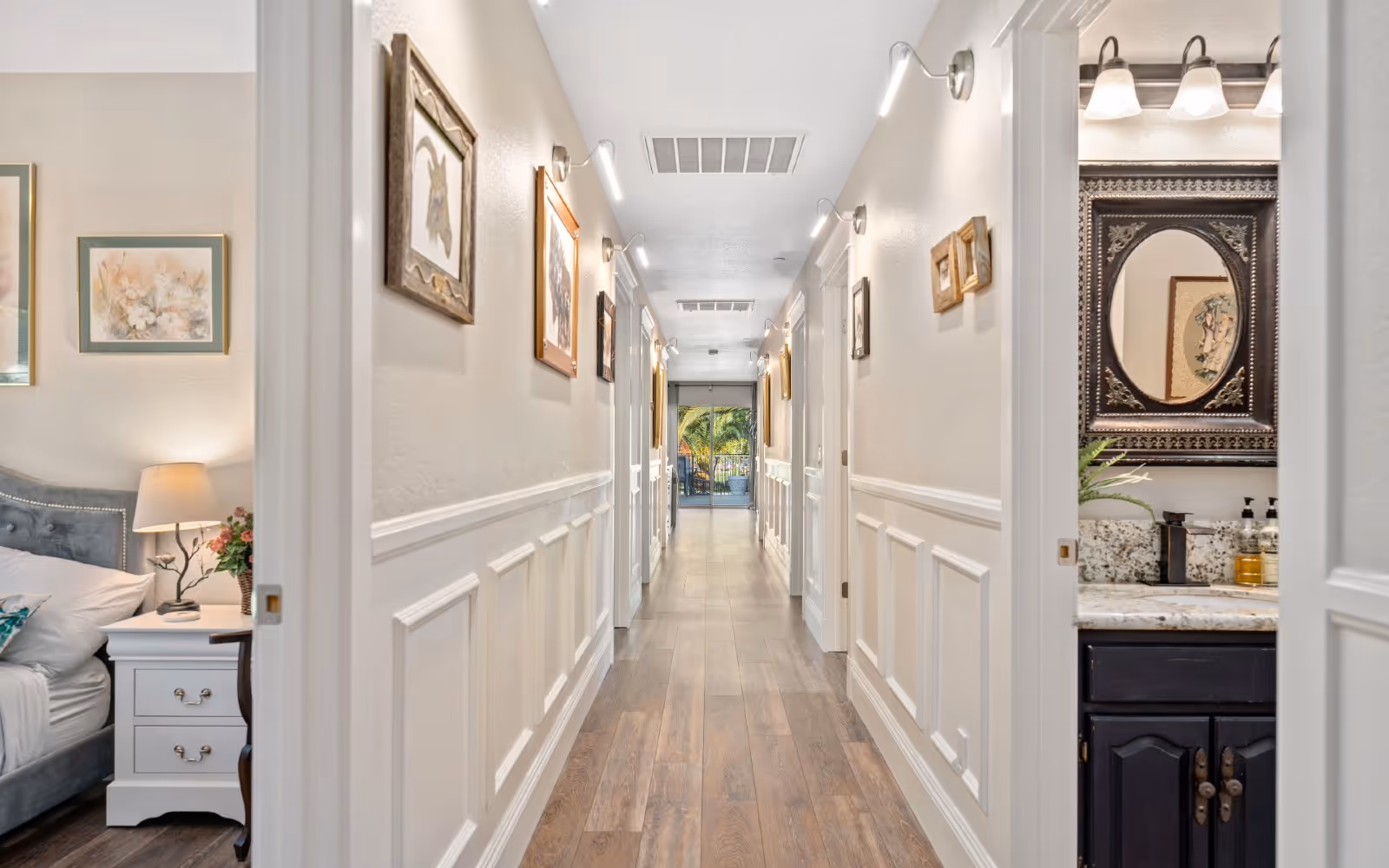 A well-lit hallway with wooden flooring and white wainscoting on the walls. The hallway is decorated with framed pictures and has multiple doors on both sides. On the left, a glimpse of a bedroom with a bed, nightstand, lamp, and floral artwork is visible. On the right, a bathroom sink with a decorative mirror and soap dispensers can be seen. At the end of the hallway, there is a glass door leading to an outdoor area with greenery.