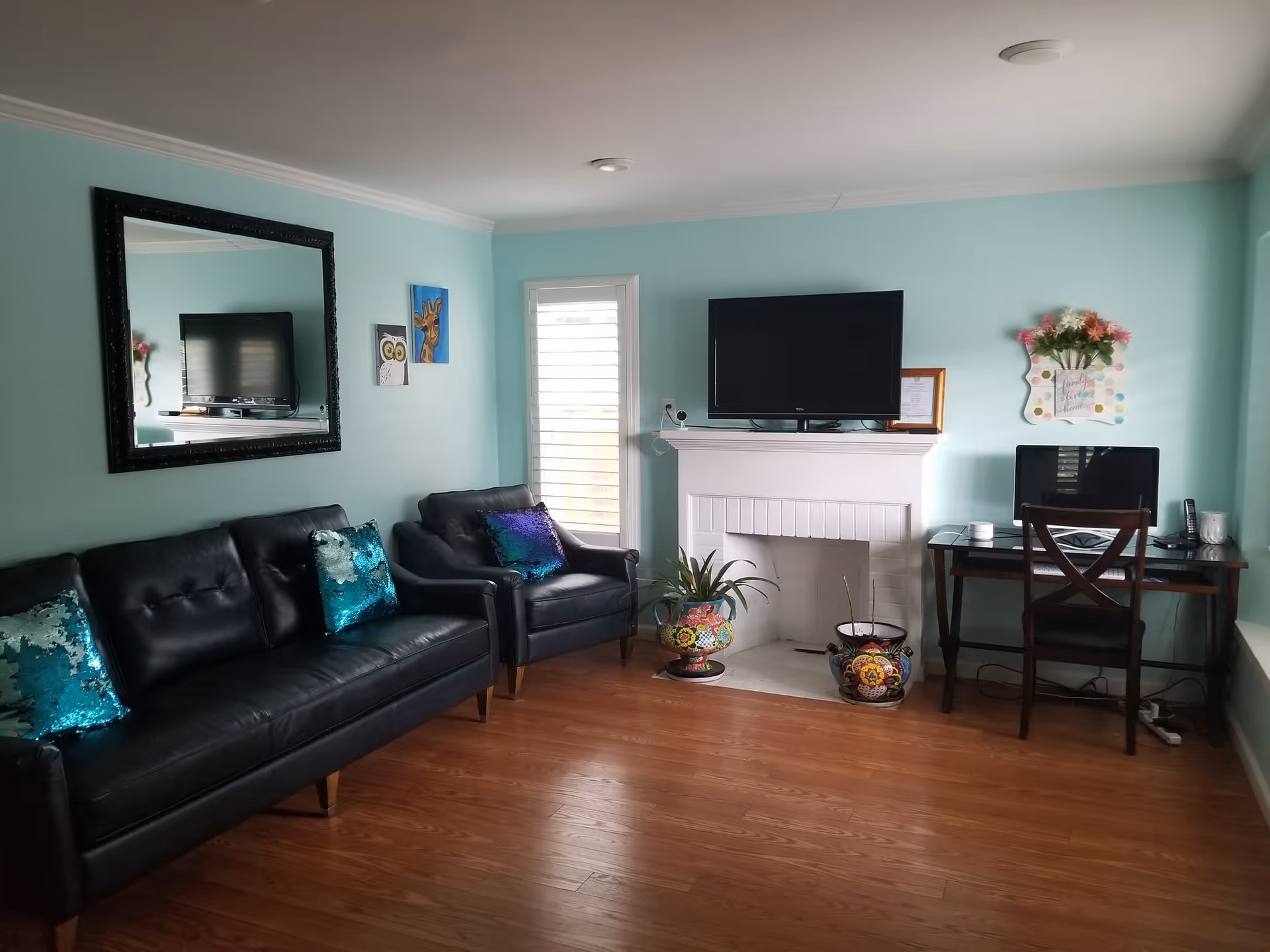 A cozy living room with light blue walls, a black leather sofa and armchair with shiny blue and purple pillows, a large mirror on the wall, a flat-screen TV mounted above a white fireplace, two colorful flower pots on the hearth, a desk with a computer and chair, and a window with white shutters.