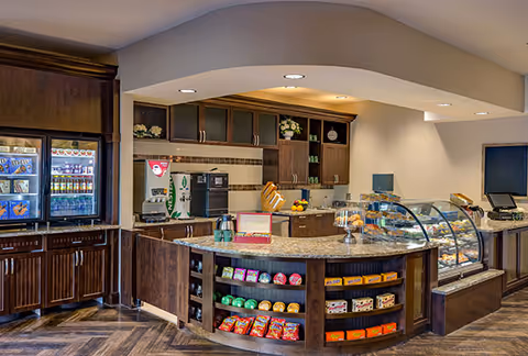 Cafe-style serving counter with glass pastry displays, snack shelves, beverage coolers, and a coffee station in a communal area.