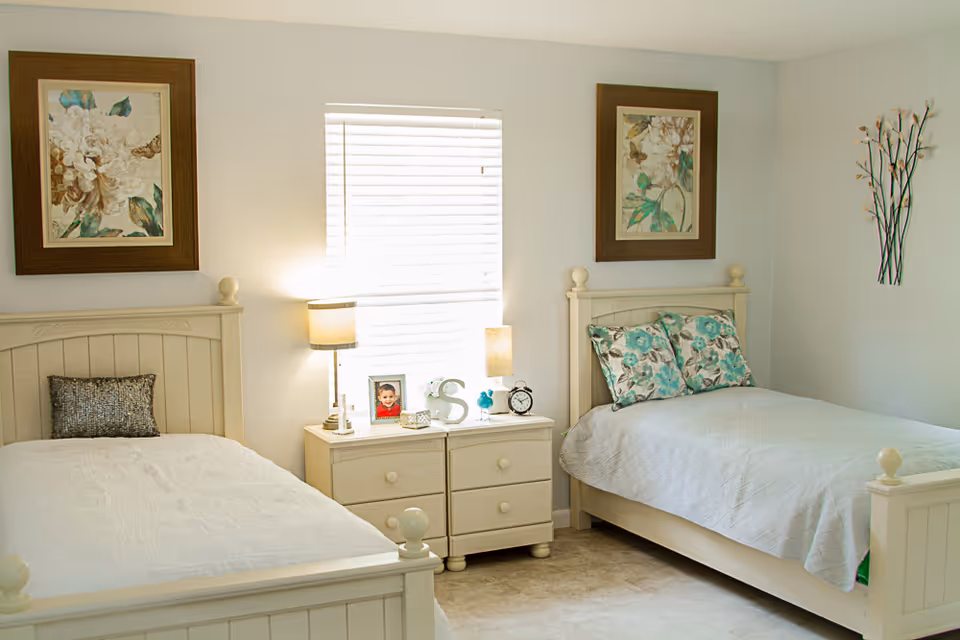 Bright bedroom with two twin beds, a shared nightstand and lamp under a central window, floral artwork on the walls.