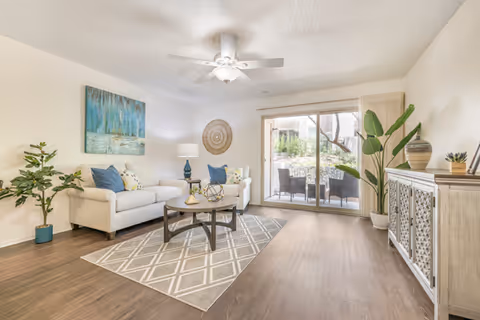 A bright living room with a white sofa adorned with blue and patterned cushions, a wooden coffee table on a gray patterned rug, a ceiling fan with light, a large potted plant, and a sliding glass door leading to an outdoor patio with chairs and a table.