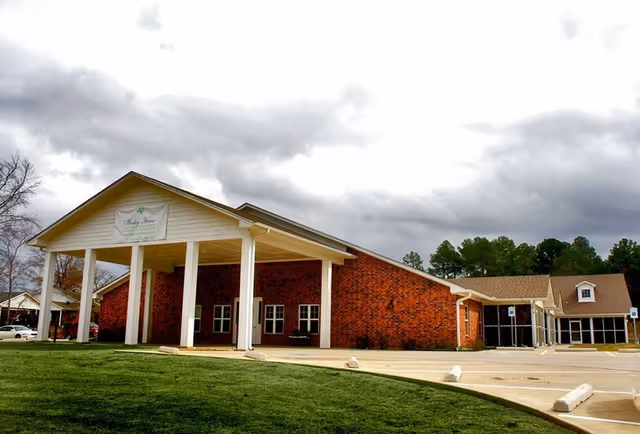Exterior view of Wesley House, a single-story brick building with a covered entrance supported by white columns. The sky is cloudy, and there is a green lawn in front of the building along with a parking area.