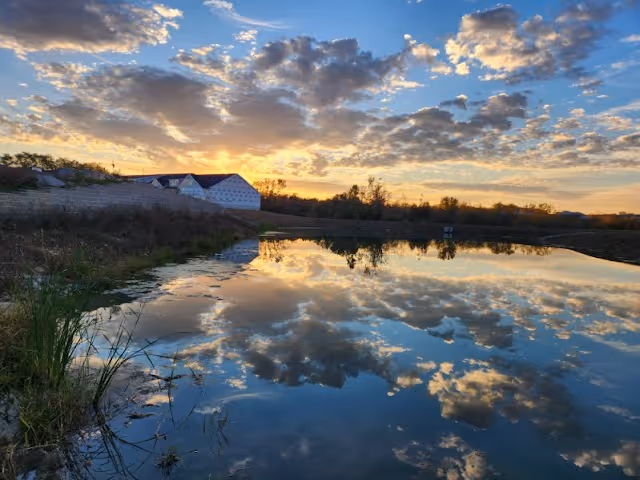 A serene outdoor scene at sunset with a calm body of water reflecting a partly cloudy sky. There is vegetation along the water's edge and a building with a red roof in the background.