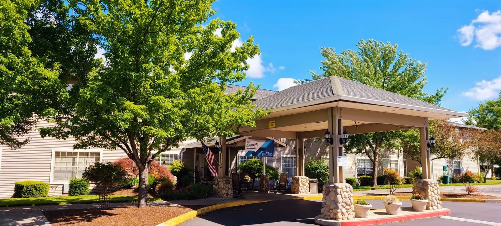 Entrance of Lakeland Senior Living facility with a covered drop-off area supported by stone pillars, surrounded by green trees and shrubs under a blue sky with some clouds.