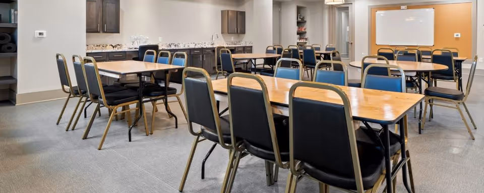 Multipurpose community dining room with rows of tables and chairs, a kitchenette along the back wall, and a whiteboard.