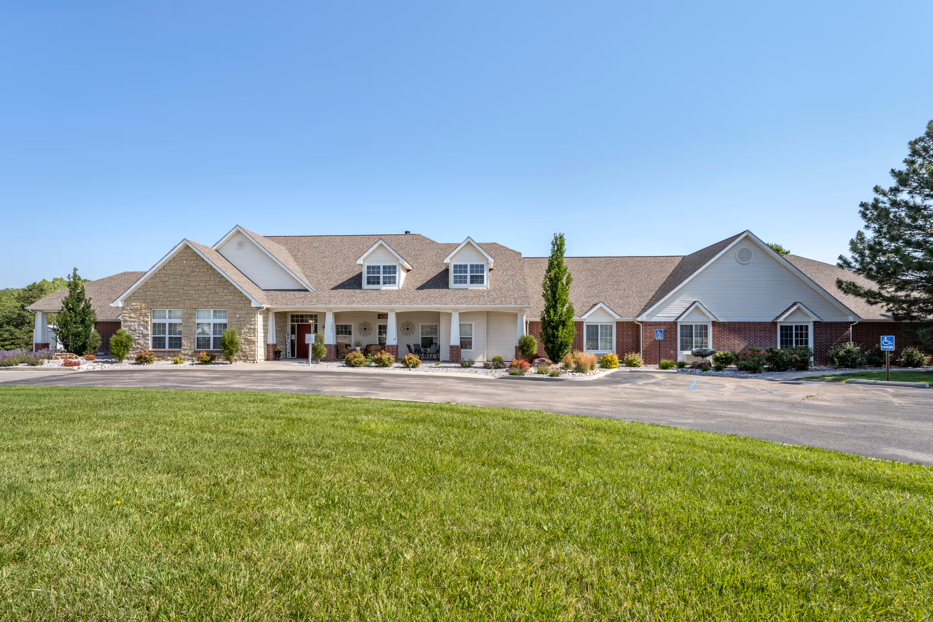Front exterior view of a single-story senior living facility building with a combination of brick and stone facade, multiple windows, a covered entrance with columns, surrounded by well-maintained landscaping and a green lawn under a clear blue sky.