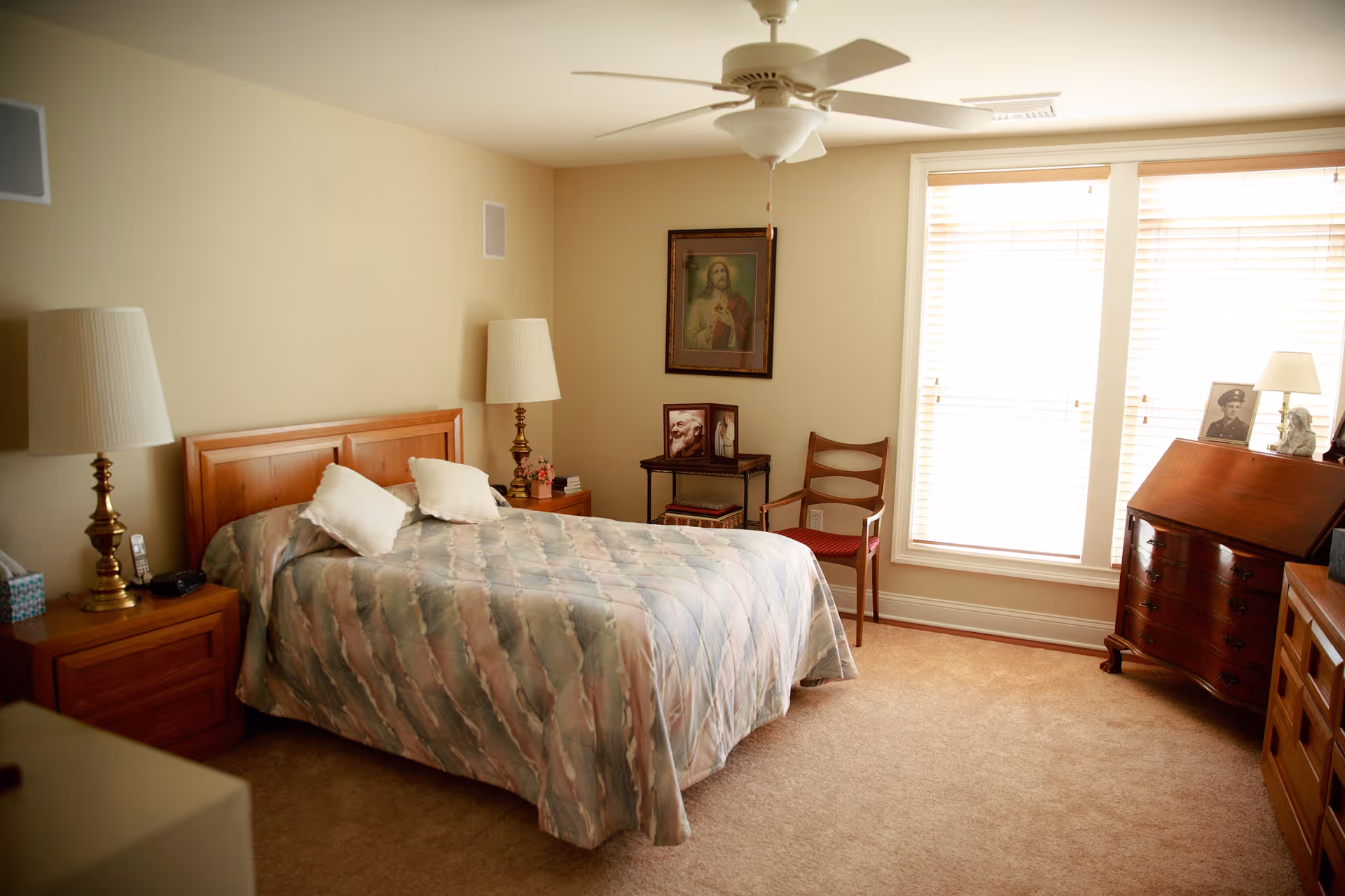A cozy bedroom with a double bed covered in a patterned quilt and two white pillows. There are two wooden nightstands with lamps on either side of the bed. A ceiling fan is mounted above. The room has beige walls and carpeted floor. A framed picture of Jesus hangs on the wall above a small table with framed photos and a chair next to it. Large windows with blinds allow natural light to fill the room. A wooden dresser with more framed photos and a lamp is positioned near the windows.