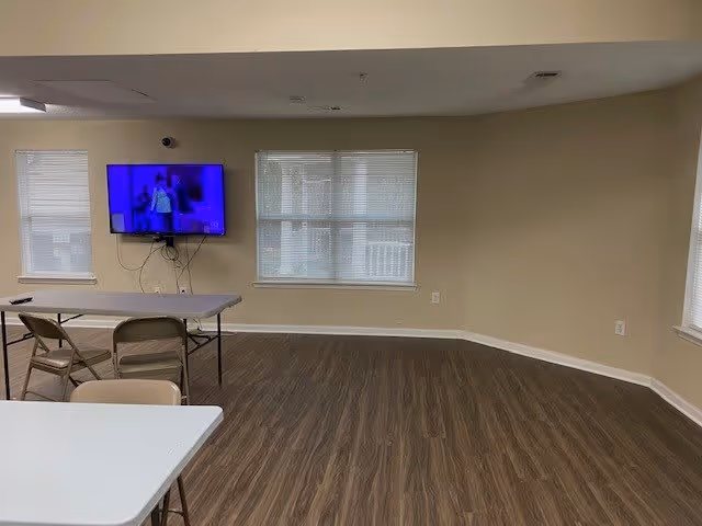 Empty common room with folding tables and chairs, a wall-mounted TV, and windows.